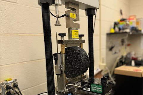 Metal jig cradling an asphalt specimen and shown in a traditional load frame in an asphalt laboratory